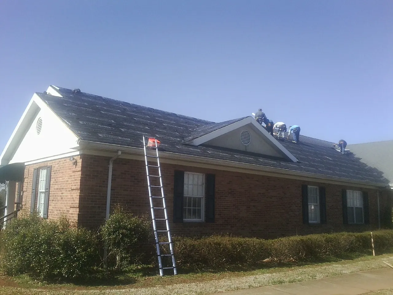 Crew tearing shingles off a brick ranch roof with a ladder set — Emergency Roof Repair work by our crew in Mays