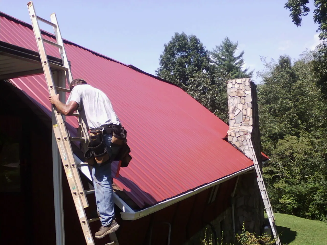 Roofer on a ladder beside a red metal roof and stone chimney — Soffit & Fascia Repair by Roofing Brothers in Mays